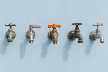 Five different outdoor water taps in a row on a pale blue wall, the middle one with an orange handle.
